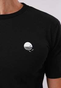 T-shirt noir avec un petit motif blanc représentant un soleil se couchant sur l'eau et trois oiseaux volant près de la zone de la poitrine.