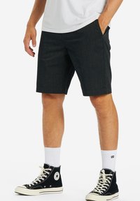 Uomo in pantaloni corti neri, calze bianche a girocollo, sneakers alte nere Converse e una maglietta bianca, con una mano in tasca.