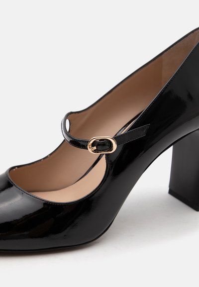 Chaussure Mary Jane en cuir verni noir avec boucle dorée et talon bloc de hauteur moyenne sur fond blanc.
