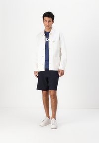 Bela bombažna jakna z zapiranjem na gumb, dvema sprednjima žepoma, v kombinaciji z modrim T-shirtom, mornarsko modrimi kratkimi hlačami in belimi supergami.
