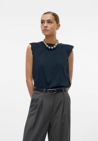 Marineblauw mouwloos topje met kantaccenten op de schouders, gecombineerd met grijze geplooide broek en een stevige zilveren ketting.