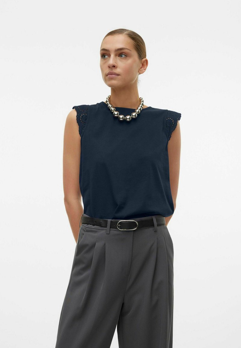 Marineblauw mouwloos topje met kantaccenten op de schouders, gecombineerd met grijze geplooide broek en een stevige zilveren ketting.