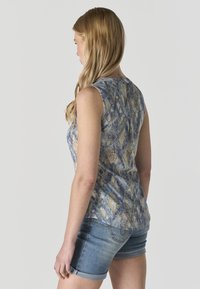 Mujer con cabello rubio largo, vistiendo una blusa sin mangas con estampado azul y beige y pantalones cortos de mezclilla clara, de espaldas y mirando hacia un lado.
