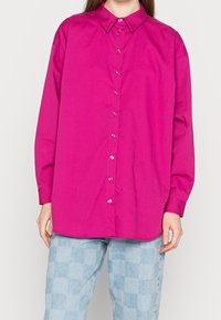 Kvinna klädd i en lysande rosa skjorta med långa ärmar och ljusblå rutiga jeans, stående mot en ljus vit bakgrund.