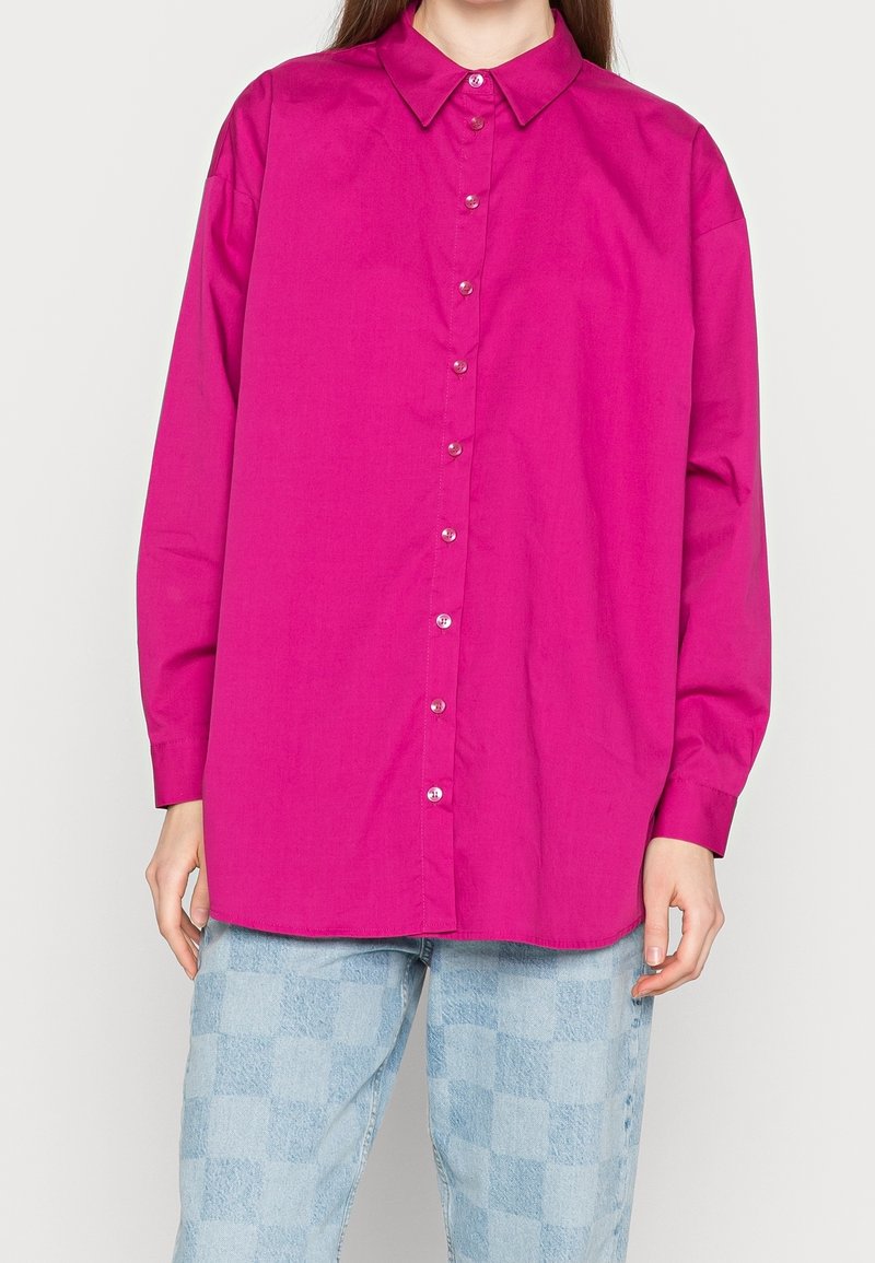 Kvinna klädd i en lysande rosa skjorta med långa ärmar och ljusblå rutiga jeans, stående mot en ljus vit bakgrund.