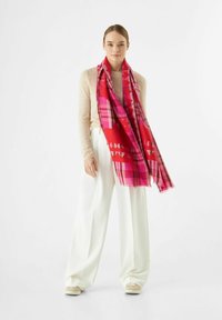 Jeune femme portant un pull beige, un pantalon large blanc, des chaussures beiges et une grande écharpe à motifs rouges et roses, se tenant devant un fond blanc.