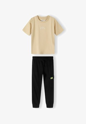 Tricou din bumbac bej, cu text brodat, asortat cu pantaloni jogger negri, având un elastic la talie și un buzunar lateral cu accente verzi.