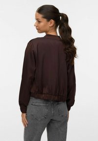Giacca bomber in satin marrone con polsini e orlo a costine, caratterizzata da una texture liscia e un taglio corto, abbinata a jeans grigi.