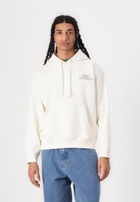 Cream-colored hoodie with a front pocket, drawstrings, and "OBEY STUDIOS" printed on the left chest. Paired with light blue jeans.