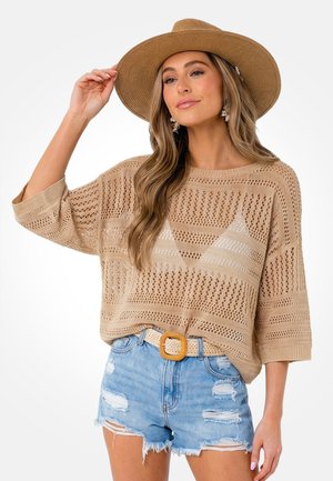 Frau mit breitkrempigem Strohhut, beigefarbenem Strickpullover, beigem Gürtel und zerrissenen hellblauen Jeansshorts, posiert vor weißem Hintergrund.