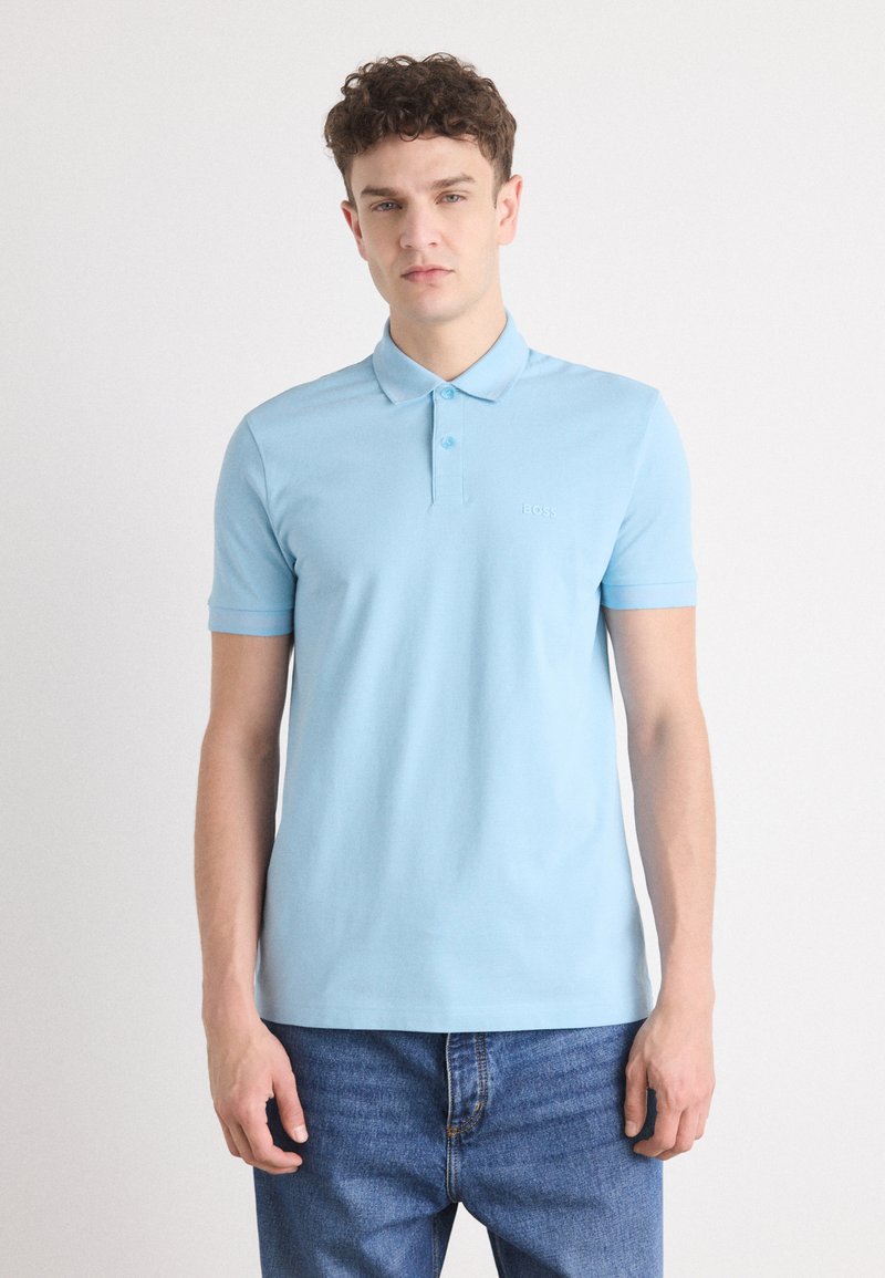 Gaiši zils kokvilnas polo krekls ar klasisku apkakli, īsām piedurknēm un smalku logo izšuvumu uz krūšu. Uzvilkts ar tumšiem džinsa biksēm.