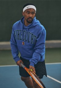 Sweatshirt azul com "MIKECOURT" em letras em negrito, combinado com calções pretos. Segurando uma raquete de ténis num campo com um fundo verde.