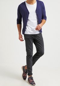 Marinblå cardigan över en vit T-shirt, i kombination med mörka slim-fit jeans och flerfärgade mönstrade sneakers. Enkel design med knappdetaljer.