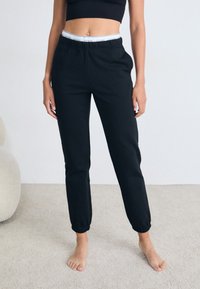 Pantaloni della tuta neri con un design affusolato, elastico in vita con un logo bianco e polsini a coste. Realizzati in tessuto morbido, indossati a piedi nudi.