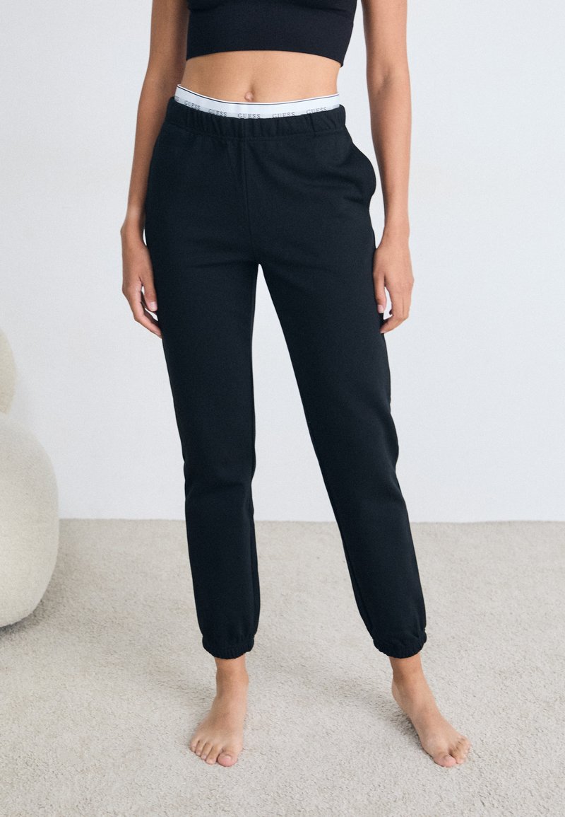 Pantaloni della tuta neri con un design affusolato, elastico in vita con un logo bianco e polsini a coste. Realizzati in tessuto morbido, indossati a piedi nudi.