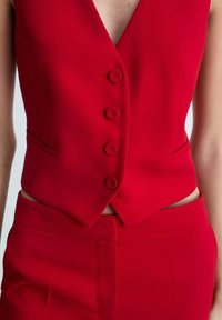 Rotes maßgeschneidertes Weste mit V-Ausschnitt, fünf passenden Knöpfen und Seitentaschen, kombiniert mit roten hochtaillierten Hosen mit einem Schlitzdetail.