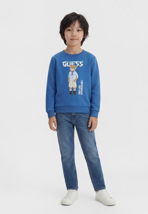 Ragazzo giovane in piedi di fronte, indossa una felpa blu con grafica dell’orso, jeans blu e scarpe da ginnastica bianche su sfondo semplice.