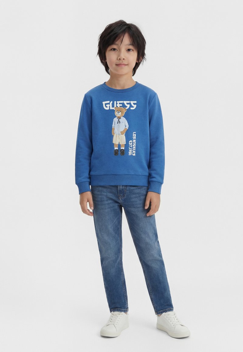 Ένα μικρό αγόρι στέκεται μπροστά, φοράει μπλε φούτερ με γραφικό αρκούδας, μπλε τζιν και λευκά αθλητικά παπούτσια σε απλό φόντο.