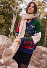 Pleteni višebojni džemper s kombinacijom uzoraka i tekstura, pretežno u mornarsko plavoj, zelenoj, crvenoj i bijeloj boji, uparen s kremastom maramom.