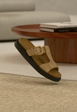 Sandale en daim beige avec boucle dorée et semelle noire flottant au-dessus d'un tapis beige dans un salon moderne avec une table basse en bois et des livres.