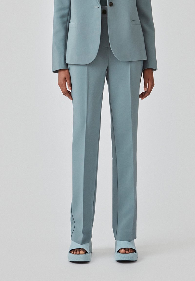 Personne portant un pantalon de costume bleu clair sur mesure et des chaussures à plateforme ouvertes assorties, debout devant un fond uni.