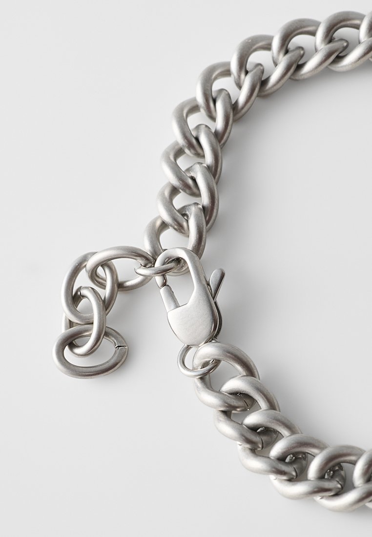 Cadena de metal plateado con grandes eslabones entrelazados, que cuenta con un cierre de resorte y un acabado liso y pulido. El diseño texturizado resalta la durabilidad.