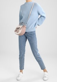 Personne portant un pull bleu clair, un jean bleu, des baskets blanches, tenant un sac à bandoulière rose et blanc avec un détail nœud.