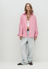 Camicia oversize a righe rosa con bottoni, top corto bianco sotto, jeans a gamba larga blu chiaro, ballerine marrone scuro, collane di argento a strati.