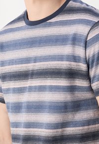 Homme portant un t-shirt en maille à rayures bleues, blanches et roses, à manches courtes et col rond bleu foncé, torse et cou visibles.