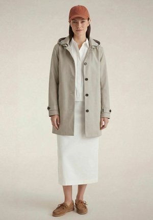Donna che indossa un cappotto beige con cappuccio, camicia bianca e gonna, scarpe marroni e cappello color ruggine, in piedi davanti a uno sfondo neutro.