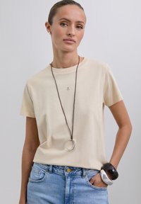 Camiseta de manga corta beige hecha de algodón suave; presenta un pequeño detalle de logo en la parte delantera. Combinada con jeans de mezclilla azul claro.