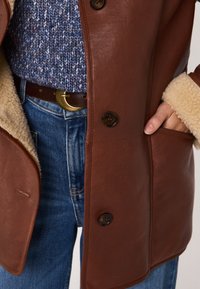 Manteau en cuir marron avec un col en peau de mouton, doté de boutons foncés, d'une poche latérale, et d'un pull en maille bleu en dessous.
