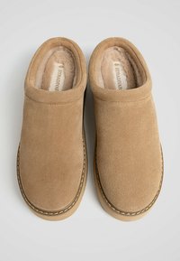 Tan suede clogs with a soft, plush interior lining. Rounded toe shape and stitched detailing along the sole. Adjustable fit without closure.