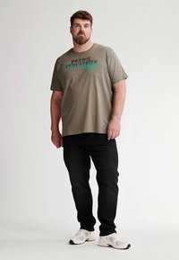 Olivově zelené bavlněné tričko s černým a tyrkysovým motivem "PETROL INDUSTRIES", doplněné černými džíny a bílými sportovními botami.