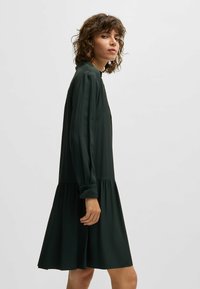 Frau mit lockigem Haar in einem dunkelgrünen Langarmkleid, die seitlich vor einem schlichten hellen Hintergrund steht.
