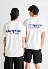 White cotton t-shirts feature "tommy jeans" logo in multicolored gradient on the back. Worn with black shorts, displayed from behind.