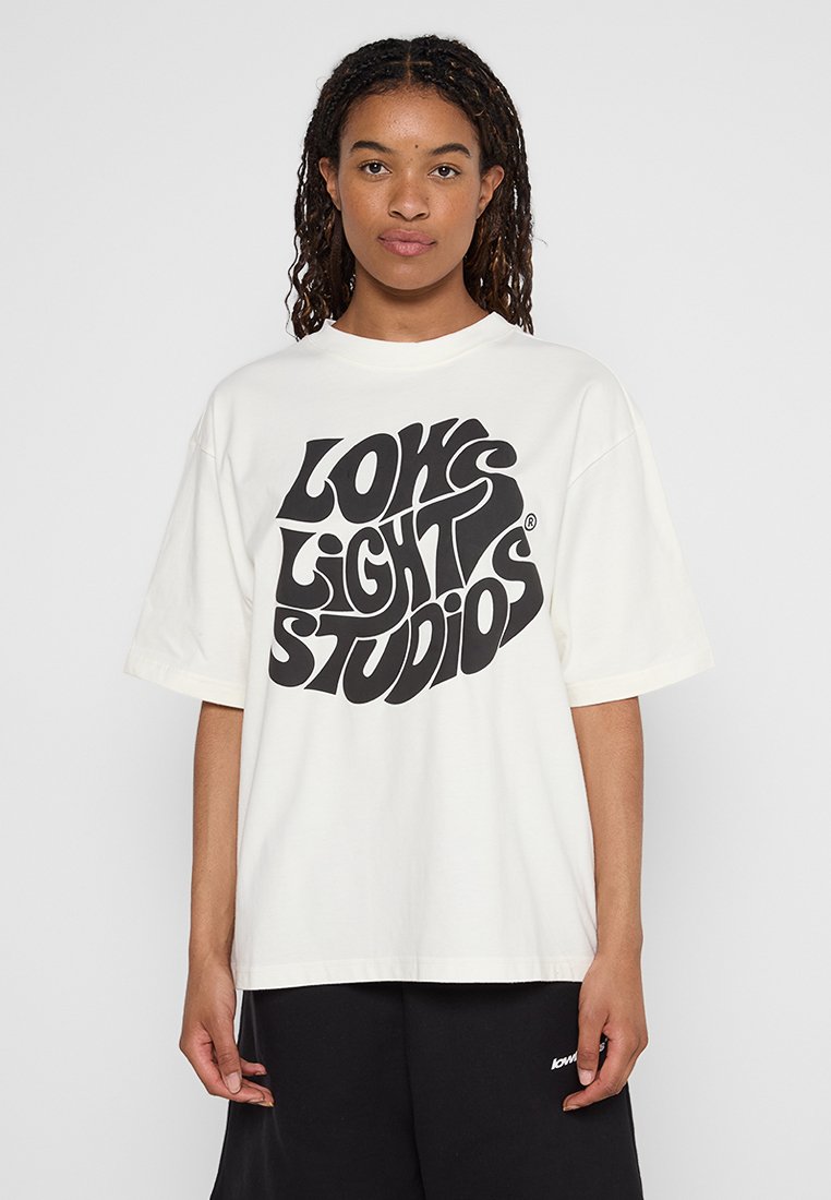 T-shirt en coton crème avec une coupe oversized, col rond et texte graphique noir "LOW LIGHT STUDIOS" affiché en avant.