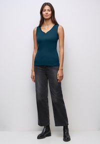 Top senza maniche di colore teal con décolleté a V e piccoli bottoni, abbinato a jeans a gamba larga grigio scuro e stivaletti neri.