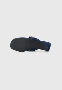 Sandal à talons hauts en denim avec une tige bleu foncé, une fine bride autour de la cheville, des accents en métal doré et une semelle noire texturée.