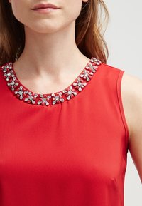 Top rosso senza maniche caratterizzato da un tessuto texturizzato con un design floreale di perline attorno al colletto, che aggiunge arricchimenti e dettagli.
