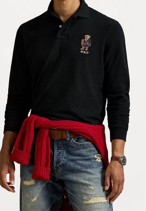 Homme portant un polo noir à manches longues avec un logo d'ours brodé, un jean bleu usé, un pull rouge noué à la taille et une montre-bracelet.