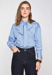Blusa a righe blu e bianche con un grande fiocco, maniche lunghe e polsini con bottoni, abbinata a jeans scuri a vita alta.