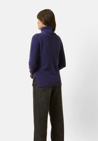 Fialový pletený svetr s vysokým límcem, bočními rozparky a žebrovaným lemem. V kombinaci s tmavými, volnými kalhotami, pohled zezadu.