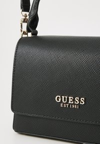 Juodas tekstūruotas rankinys su auksiniu "GUESS EST 1981" logotipu. Yra struktūruota forma ir nuimama petneša. Išskirtinis dizainas.