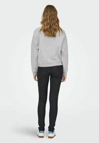 Kvinde med bølget lysebrunt hår står med ryggen til, iført en grå sweatshirt, sorte skinny jeans og hvide sneakers med blå accenter.