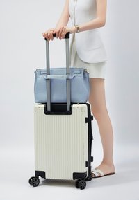 Valigia rigida bianca con scanalature verticali, dettagli neri e quattro ruote, sovrastata da una borsa in pelle azzurro chiaro.