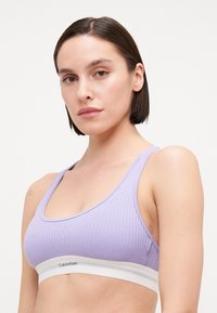 Levanduľový rebrovaný bralet s širokým bielym pásom značky Calvin Klein, ktorý má výstrih do tvaru lopatky a hrubé ramienka. Hladká textúra, ležérny dizajn.
