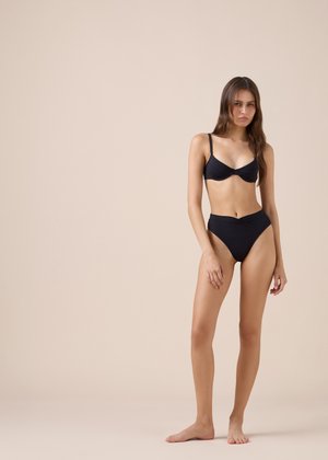 Kobieta stoi boso na beżowej podłodze w czarnym bikini na jednolitym beżowym tle, patrząc na kamerę z neutralnym wyrazem twarzy.