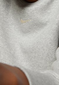 Šedá mikina vyrobená z měkké tkaniny, s malým reflexním béžovým logem Nike na hrudi a žebrovanými manžetami.