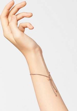 Bracelet en or rose avec un design de chaîne délicate et deux extrémités pendantes, sécurisé par un fermoir circulaire. Texture lisse et polie.
